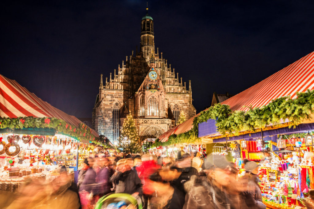 Huge crowd of people moving over Nuremberg´s world-famous christmas market (Christkindlsmarkt) at night, passing colorful illuminated christmas decoration and food stalls. Nuremberg´s landmark Frauenkirche (Church of our Lady) can be seen in the back.